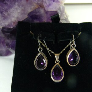 May include: A silver jewelry set featuring a teardrop-shaped amethyst pendant necklace and matching earrings. The deep purple gemstones are set in silver bezels, creating a striking contrast. The set is displayed on a black velvet surface.