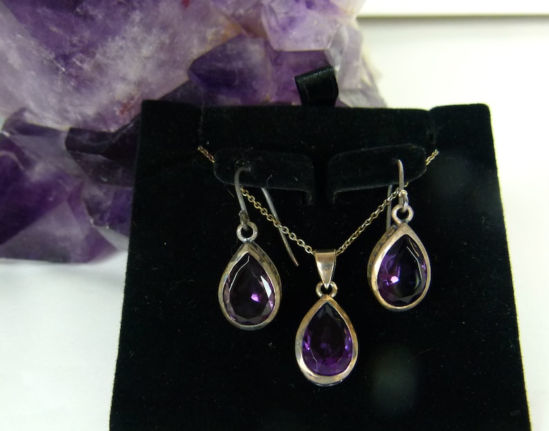 May include: A silver jewelry set featuring a teardrop-shaped amethyst pendant necklace and matching earrings. The deep purple gemstones are set in silver bezels, creating a striking contrast. The set is displayed on a black velvet surface.