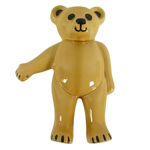 May include: A ceramic teddy bear figurine in a standing pose. The bear is a light tan color with black accents on the eyes, nose, and paws. The bear's arm is extended, and it has a glossy finish.