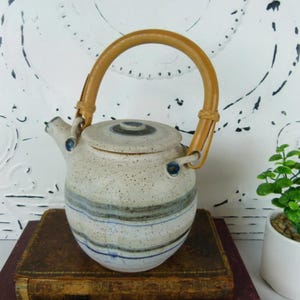 May include: A ceramic teapot with a speckled, off-white body and a bamboo handle. The teapot features blue and gray horizontal stripes and a matching lid. The handle is arched and attached with woven detailing. A small spout and blue accents complete the design.
