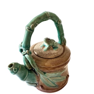 May include: A ceramic teapot with a green bamboo-style handle and spout. The body is brown with green accents, including a frog on the lid and leaf details. The teapot is designed with a unique, artistic flair.