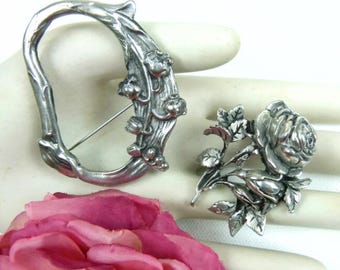 May include: Two silver tone brooches featuring floral designs. One brooch is a stylized branch with flowers and leaves. The other brooch is a single rose with leaves.