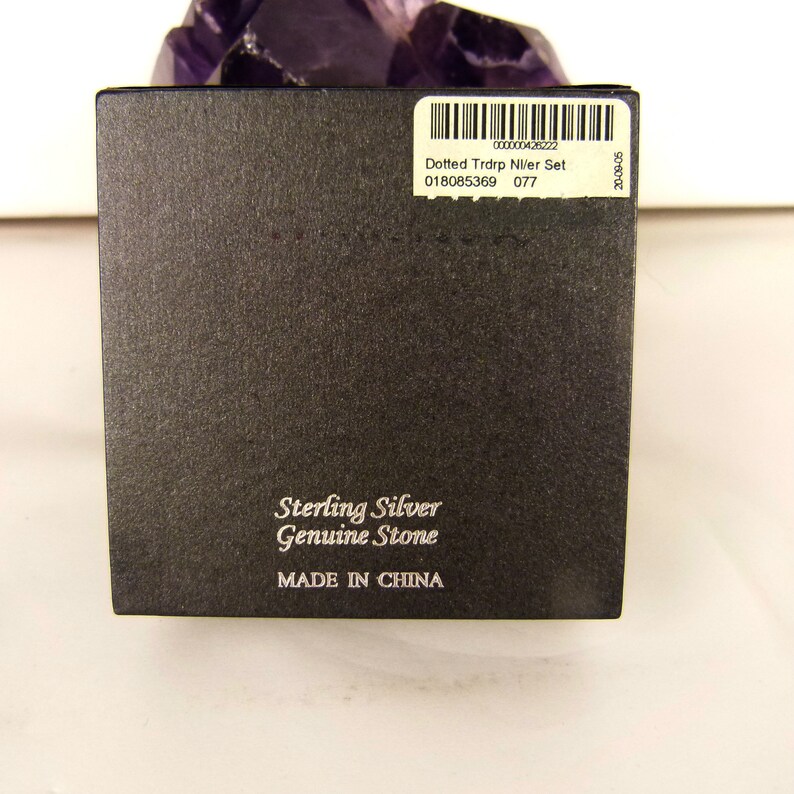 May include: A black box with the text "Sterling Silver Genuine Stone" and "MADE IN CHINA". A sticker on the box reads "Dotted Trdrp NI/er Set". The box is set against a white background and a purple crystal.