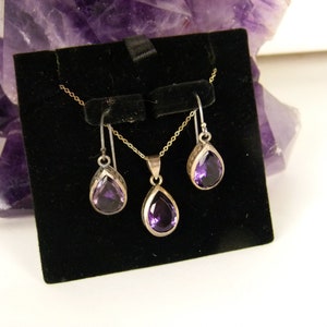 May include: A jewelry set featuring a silver necklace and matching earrings, each with a teardrop-shaped, deep purple gemstone. The earrings hang from silver hooks, and the pendant hangs from a delicate chain. The set is displayed on a black velvet background.