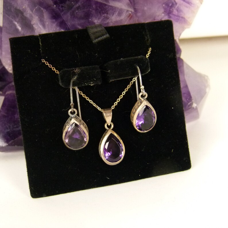 May include: A jewelry set featuring a silver necklace and matching earrings, each with a teardrop-shaped, deep purple gemstone. The earrings hang from silver hooks, and the pendant hangs from a delicate chain. The set is displayed on a black velvet background.