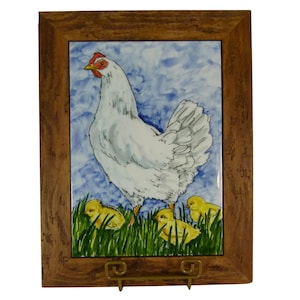 May include: A rectangular ceramic tile featuring a white hen with a red comb standing in green grass with three yellow chicks, set against a blue sky background. The tile is encased in a brown wooden frame and rests on a gold metal stand.