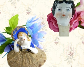 May include: Two antique porcelain doll heads and a doll pin cushion. One doll head has black hair and a painted face. The other is a doll with a blue hat and a brown skirt. The pin cushion has a brown mesh skirt.