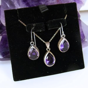 May include: A jewelry set featuring a silver necklace with a teardrop-shaped amethyst pendant and matching earrings. The earrings and pendant are displayed on a black velvet background. The set is presented on a black display stand.