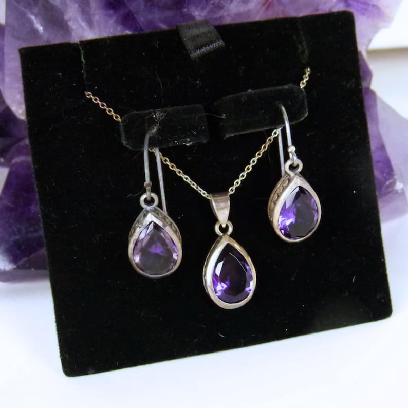 May include: A jewelry set featuring a silver necklace with a teardrop-shaped amethyst pendant and matching earrings. The earrings and pendant are displayed on a black velvet background. The set is presented on a black display stand.