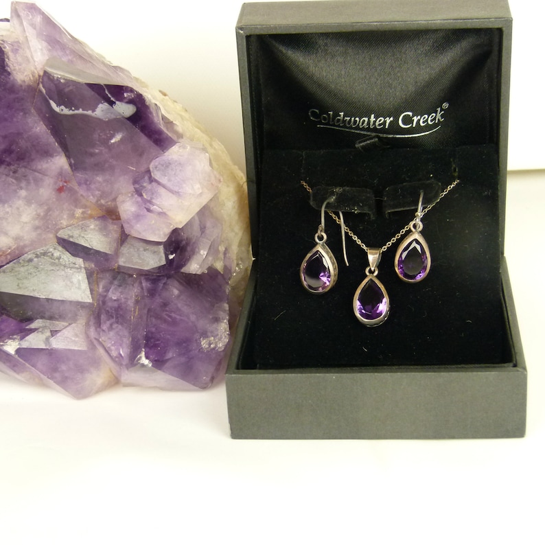 May include: A silver necklace and earrings set with teardrop-shaped purple gemstones, displayed in a black box. The box is open, revealing the jewelry against a black velvet interior. A large, raw amethyst crystal is in the background.