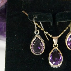 May include: A jewelry set featuring a silver necklace with a teardrop-shaped purple gemstone pendant, and matching earrings. The gemstones are faceted and set in silver bezels. The set is displayed on a black surface.