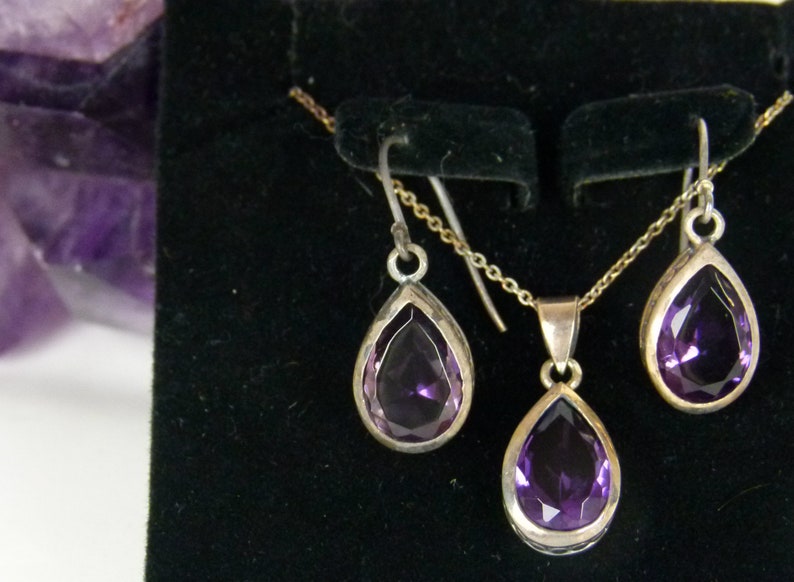 May include: A jewelry set featuring a silver necklace with a teardrop-shaped purple gemstone pendant, and matching earrings. The gemstones are faceted and set in silver bezels. The set is displayed on a black surface.