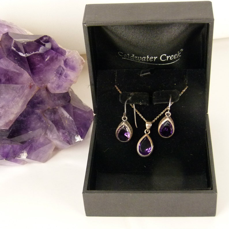 May include: A jewelry set featuring a necklace and earrings with teardrop-shaped, deep purple gemstones. The set is displayed in a black box with the brand name "Coldwater Creek" on the lid. A large, raw amethyst crystal is visible in the background.
