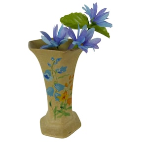 May include: A beige, vintage-style vase with a flared top and square base, adorned with hand-painted blue and yellow floral designs. The vase holds several blue artificial flowers and a green leaf, creating a delicate and charming display.