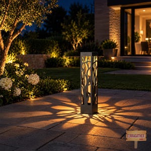 May include: A rectangular outdoor lantern with a dark metal frame featuring a laser-cut tree branch pattern. The lantern emits a warm, golden light that projects a radial, star-like shadow pattern onto the stone patio surface below it.