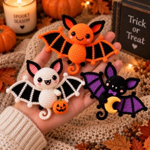 May include: Three crocheted bat ornaments featuring distinct color schemes: one orange and black, one white and black holding a small orange pumpkin, and one black and purple holding a yellow crescent moon. Each bat has black eyes and ears.