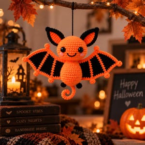 May include: A crocheted bat ornament featuring a bright orange body and head with black wing accents, large black eyes, and a small curled tail. The bat is suspended by a black string and displays a smiling face with pink cheeks.