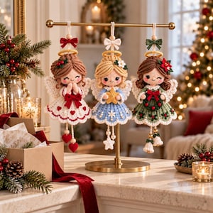 May include: Three crocheted angel ornaments hanging from a gold metal stand. The left angel wears a white dress with red hearts, the center angel a blue dress with a yellow star, and the right angel a green dress with a wreath and red bow.