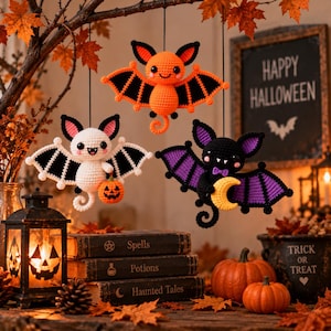 May include: Three hanging crochet bat ornaments in orange, white, and black. The orange bat has black wing accents. The white bat holds a small orange pumpkin. The black bat features purple wing details and holds a yellow crescent moon.