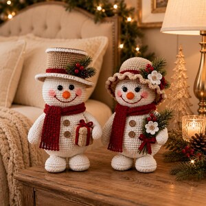 May include: Two crocheted snowmen figures with white bodies, orange carrot noses, and pink cheeks. One wears a tan top hat, the other a tan sun hat. Both have red scarves, brown buttons, and festive greenery accents with red berries and white flowers.