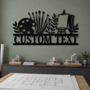 May include: A black metal wall sign featuring a silhouette of an artist's palette, several paintbrushes, leafy branches, and an easel. The lower section of the sign contains the text "CUSTOM TEXT" in a bold, capitalized, sans-serif font.