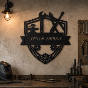 May include: A black metal wall sign shaped like a shield featuring a horizontal banner with the text SMITH FAMILY. The design includes silhouettes of a hammer, anvil, wrench, and pliers, along with decorative floral elements at the base.