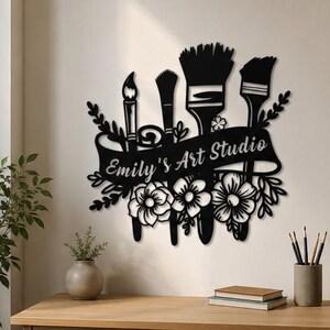 Peut inclure: Une décoration murale en métal noir représentant quatre pinceaux de tailles différentes disposés verticalement. Une bannière centrale traversant les pinceaux affiche le texte Emily's Art Studio. Le motif est orné de fleurs et de feuilles.