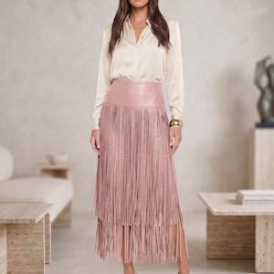 May include: A long, dusty rose-coloured leather skirt featuring two tiers of long, thin fringe that hang from a smooth, high-waisted band. The skirt is paired with a long-sleeved, champagne-coloured satin button-down shirt and open-toed beige heels.
