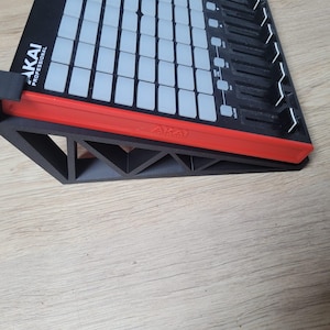 以下が含まれることがあります： 黒と赤のAkai Professional MIDIコントローラーが、黒い三角形のオープンフレームスタンドに置かれています。コントローラーには、ライトグレーの正方形パッドのグリッドと、右側に一連の垂直スライダーがあり、Akaiのロゴが確認できます。