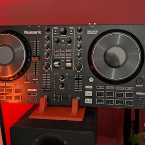 May include: A black Numark Mixtrack Platinum FX four-deck DJ controller featuring two large jog wheels, a central mixer section with volume faders, EQ knobs, performance pads, and Serato branding. The device is mounted on a red stand.