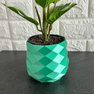 Op de afbeelding: Een levendige mintgroene plantenpot met een geometrische, gefacetteerde, diamantachtige oppervlaktestructuur. De pot bevat een groene bladplant met donkerbruine aarde zichtbaar aan de bovenkant. De structuur heeft herhalende driehoeksvormen.