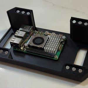 Puede incluir: Un marco de montaje negro impreso en 3D con cuatro pilares en las esquinas, cada uno con tres imanes circulares plateados incrustados. Una placa de circuito verde con un ventilador central y un disipador térmico está fijada en el marco.