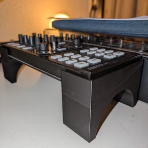 Puede incluir: Un soporte de plástico negro que sostiene un controlador de DJ negro con botones blancos y perillas negras. El controlador presenta el texto KONTROL X1 impreso en la superficie superior. El soporte tiene recortes arqueados en los laterales.