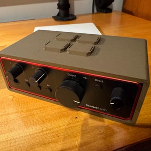 May include: A Scarlett Solo audio interface housed in a matte olive green custom enclosure. The front panel is black with red trim, featuring several black control knobs, a large central volume dial, and the text Scarlett Solo, Output, and Direct.