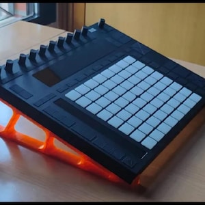 Op de afbeelding: Een zwarte muziekproductiecontroller met een raster van witte vierkante pads en een rij zwarte draaiknoppen, gemonteerd op een feloranje, schuine standaard met een open frame die het apparaat verhoogt voor beter zicht en ergonomie.
