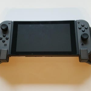 以下が含まれることがあります： 黒い画面と両側にダークグレーのコントローラーを備えた携帯型ゲーム機。デバイスの左右の端には黒い人間工学に基づいたグリップがあり、方向ボタン、ジョイスティック、アクションボタンが配置されています。