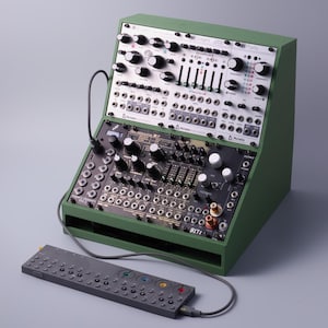 Könnte beinhalten: Ein grünes Desktop-Gehäuse für modulare Synthesizer mit mehreren silbernen und schwarzen Elektronikmodulen, Drehreglern, Schiebereglern und Patch-Punkten. Ein schmaler, dunkelgrauer, rechteckiger Controller ist per Kabel verbunden.