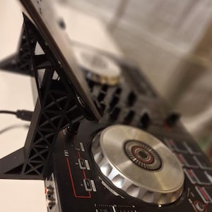 May include: A black 3D-printed plastic stand supporting a tablet device, positioned over a black DJ controller featuring silver jog wheels, various buttons, and red accent lines. The stand has a geometric lattice structure and a black power cable.