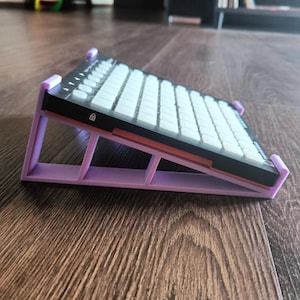 Puede incluir: Un teclado mecánico en blanco y negro sobre un soporte de plástico inclinado de color violeta claro. El teclado presenta una cuadrícula de teclas cuadradas blancas, un marco negro y un pequeño icono de candado impreso en el lateral.