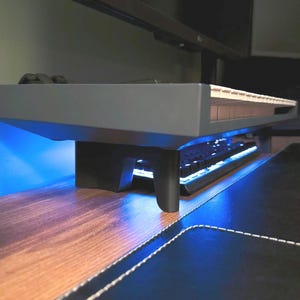 Può includere: Un supporto per monitor grigio con una solida piattaforma rialzata e due robuste gambe di supporto curve. Il supporto è posizionato sopra una tastiera meccanica nera dotata di una luminosa retroilluminazione blu, creando uno spazio libero sotto.