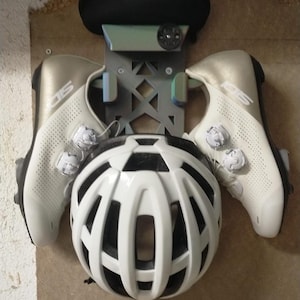 May include: A pair of white and metallic gold cycling shoes with dual dial closures, a white bicycle helmet with black interior accents, and a black saddle mounted on a grey geometric wall-mounted storage rack with a circular adjustment dial.