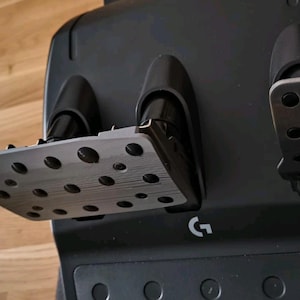 May include: A black racing pedal set featuring three metallic pedals with circular grip patterns. The base is constructed from black plastic and displays a white Logitech G logo centered at the bottom. The pedals are angled for foot operation.