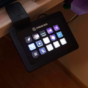 Puede incluir: Un controlador Stream Deck negro con una cuadrícula de botones cuadrados iluminados que muestran varios iconos y texto, montado en el borde de un escritorio con una abrazadera negra. El dispositivo muestra un reloj digital y accesos directos.