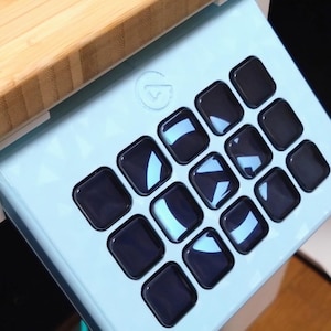 Puede incluir: Un controlador Elgato Stream Deck de color azul claro con una cuadrícula de quince teclas cuadradas de color negro brillante. El dispositivo está montado en ángulo bajo una superficie de madera clara y muestra un logotipo circular en relieve.