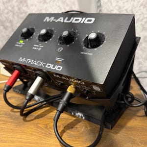 May include: A black M-Audio M-Track Duo audio interface featuring four rotary control knobs, a phantom power indicator, and multiple input and output ports. The device is mounted on a black angled stand with three connected audio cables.