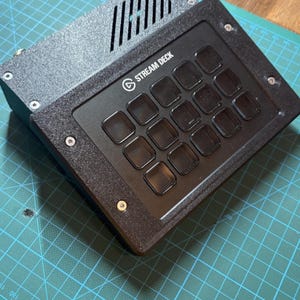 Op de afbeelding: Een zwarte Stream Deck-controller met een raster van vijftien vierkante knoppen en een behuizing met textuur. Het apparaat heeft een bovengedeelte met diagonale ventilatiesleuven en is bevestigd met zichtbare zilveren schroeven aan de randen.