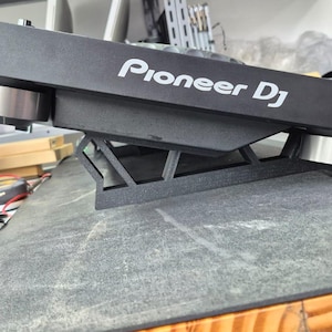 Pode incluir: Um controlador Pioneer DJ preto apoiado sobre um suporte inclinado impresso em 3D. O controlador apresenta um acabamento preto fosco com o logotipo branco da Pioneer DJ impresso na lateral. O suporte eleva o dispositivo em ângulo.