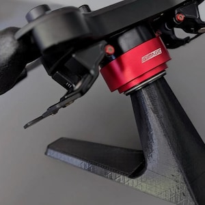 May include: A black racing steering wheel mounted on a black 3D-printed stand. The steering wheel features a central red metallic quick-release hub with the white text SIMAGIC printed on it, black textured grips, and black paddle shifters.