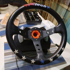 May include: A black leather racing steering wheel featuring a central orange hub, silver paddle shifters, and white and blue star embroidery along the rim. The top of the wheel displays the text R-tech in red and blue lettering with star accents.
