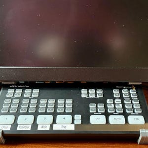 May include: A black ATEM Mini Pro video switcher featuring a control panel with numerous white buttons, including labeled keys for Close, Projector, Wide, and iPad. The device is mounted on a gray stand beneath a black computer monitor screen.